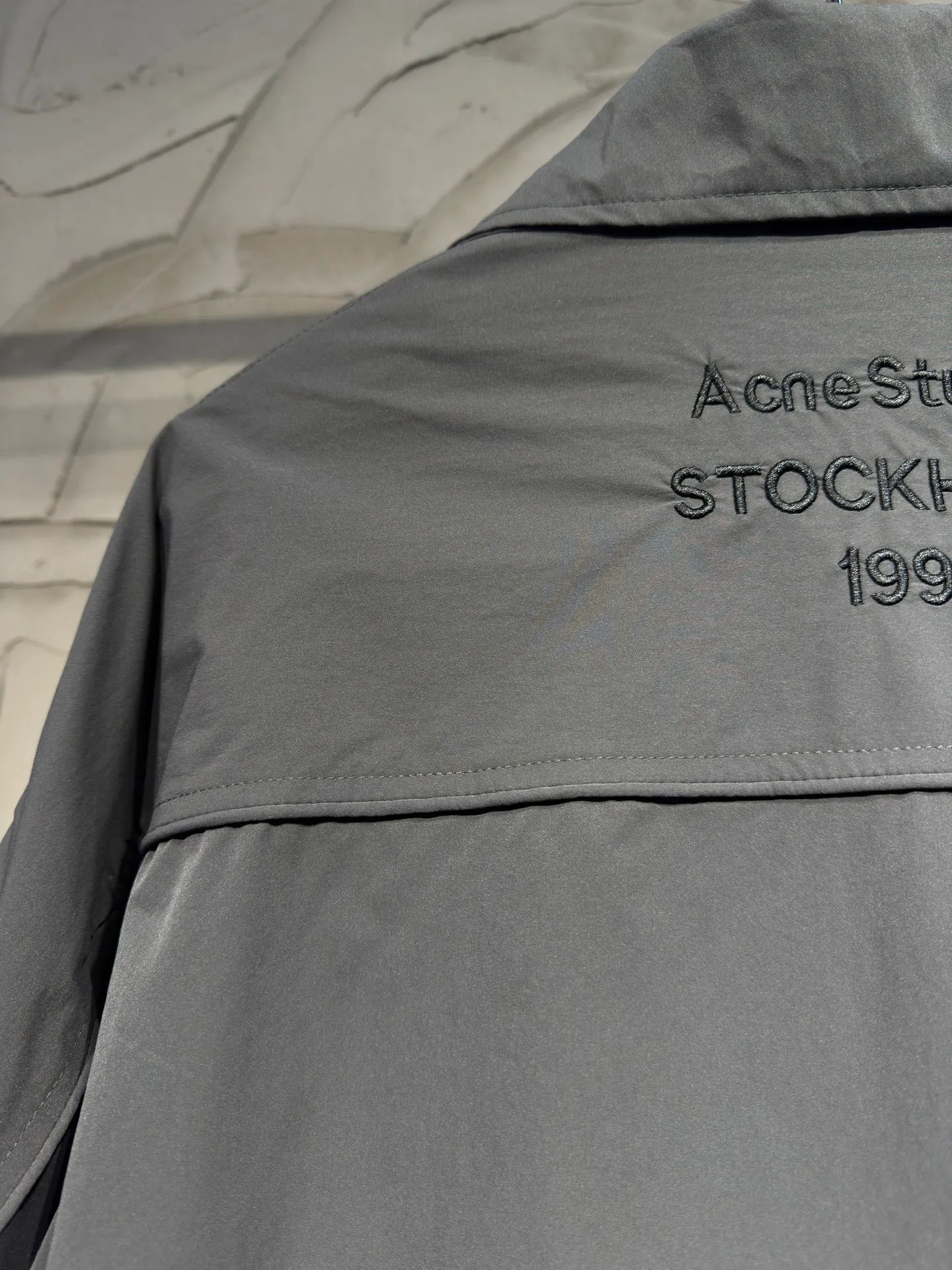 Acne Studios 26SS Large Lapel Short Jacket The fabric is made of 100% nylon (nylon/polyamide fiber) 75D high-density wrinkled nylon. The fabric has been permanently pressed and shaped by high temperature, and washed with high-pressure wrinkles at the factory to form its own irregular texture, permanently retaining the wrinkled texture, which will not disappear after washing. Matte matte coating removes the plastic glossy surface of nylon itself to create a low-key matte paper feel. Three-proof micro-coating, weak waterproof, anti-fouling, anti-static treatment, not easy to get dust. The interior is made of full-width mesh Breathable nylon lining, which reduces the stickiness of the nylon fabric to the body, improves air circulation, prevents sweating from sticking to the skin, and the zipper does not rub against the skin. The version adopts the raglan split topstitching process, and the front and rear panels are divided diagonally to expose even stitching. The three-dimensional dropped shoulders are loose and the fit is increased to increase the margin of movement. The back neckline is three-dimensional and high-density computer embroidered, and the embroidery lines are dense and not prickly. Customized all-metal matte nylon zipper, Genuine Leather embossed brand letters on the zipper leather tab, zipper tape with same color nylon fit to the body, double-locked reinforcement on both sides of the zipper to prevent pulling and tearing. Color: Gray Size: S M L